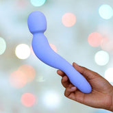 A person holds the Dame Com Wand Massager - Periwinkle against a backdrop of soft, colorful bokeh lights, highlighting the appeal of clitoral stimulation with its blue, curved design.